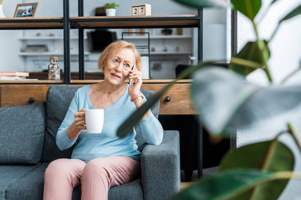 zmatený starší žena s šálkem kávy sedí na gauči a mluví na smartphone v obývacím pokoji - Fotografie, Obrázek