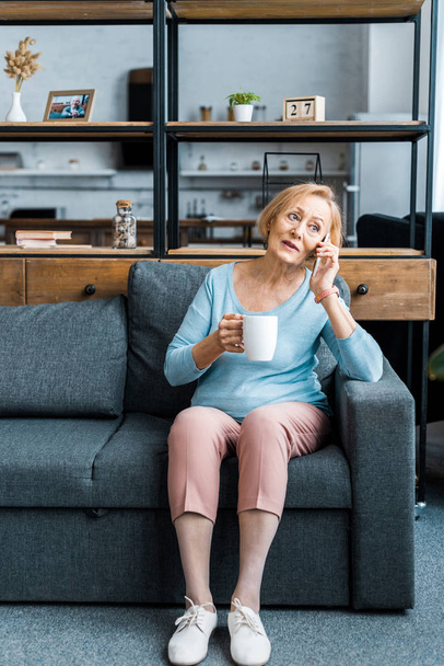 Senior vrouw met kopje koffie zittend op de Bank en praten over smartphone in woonkamer - Foto, afbeelding
