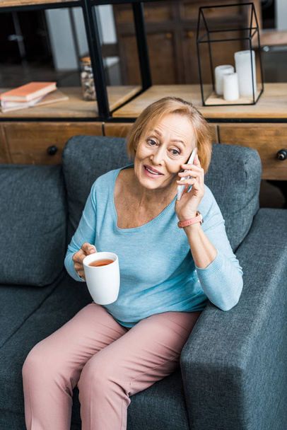Gelukkig senior vrouw met kopje koffie zittend op de Bank en praten over smartphone in woonkamer - Foto, afbeelding
