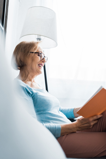επιλεκτική εστίαση του χαμογελαστό ανώτερων γυναίκα σε ποτήρια καθισμένος και ανάγνωση βιβλίων στο σπίτι  - Φωτογραφία, εικόνα