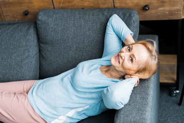 mujer mayor sonriente con las manos en la cabeza mirando a la cámara, acostada y descansando en el sofá
 - Foto, Imagen