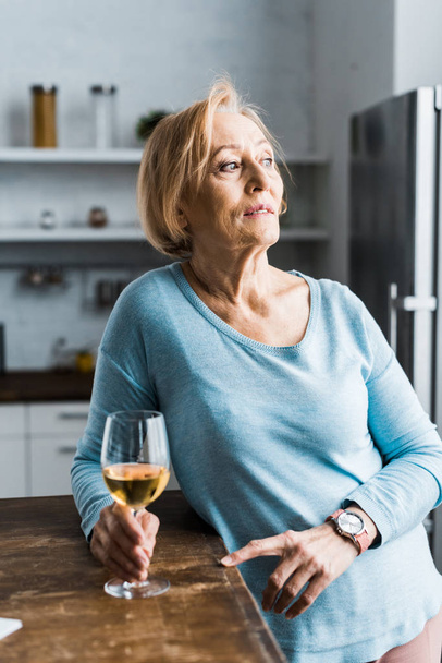 lonely senior woman holding wine glass in kitchen and looking away - Фото, зображення