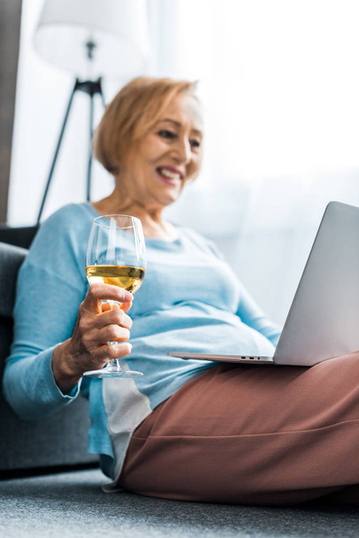 uśmiechający się starszy kobieta gospodarstwa wina szkła mając wideorozmowy na laptopa w domu - Zdjęcie, obraz