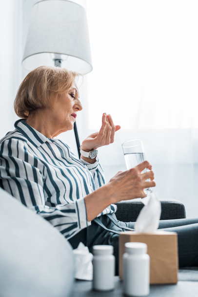 foco seletivo da mulher idosa doente tomando pílulas e segurando vidro de água em casa
 - Foto, Imagem