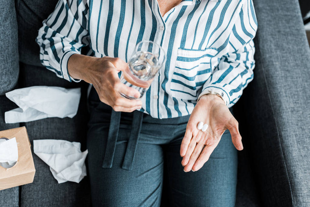 Ausgeschnittene Ansicht einer kranken Seniorin, die mit Tabletten und einem Glas Wasser zu Hause auf der Couch sitzt - Foto, Bild