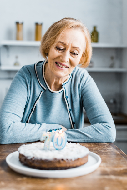 senior woman sitting at table and looking at cake with '70' sign during birthday celebration at home - Foto, Bild