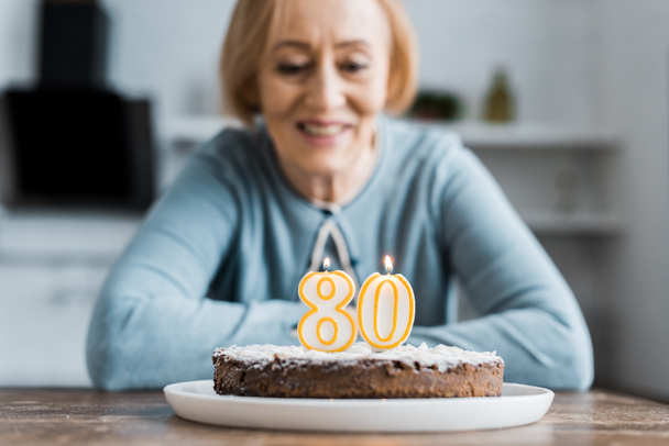 smiling senior woman sitting at table and looking at cake with '80' sign on top during birthday celebration at home - Photo, Image