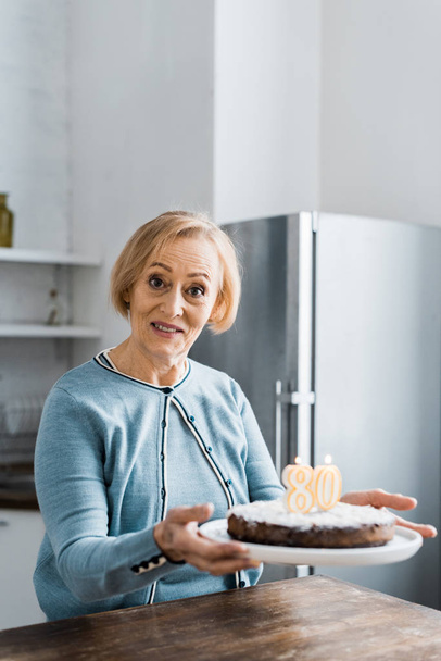 senior woman looking at camera and holding cake with '80' sign on top during birthday celebration - Foto, Bild