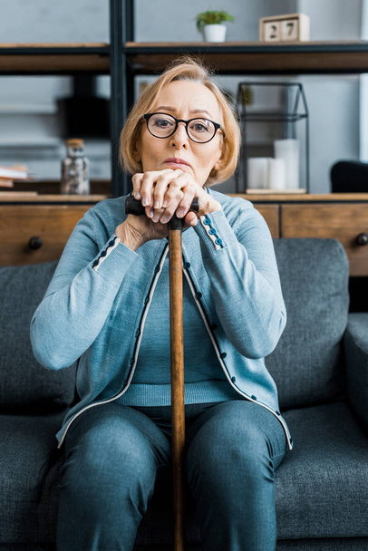 upset senior woman in glasses sitting on couch, looking at camera and leaning on walking stick in living room - Photo, Image