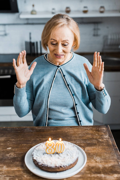 mulher sênior animado sentado e olhando para o bolo com '80' sinal no topo durante a celebração de aniversário
 - Foto, Imagem