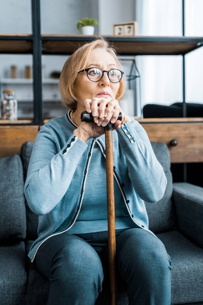 upset senior woman in glasses sitting on couch and leaning on walking stick in living room - Photo, Image
