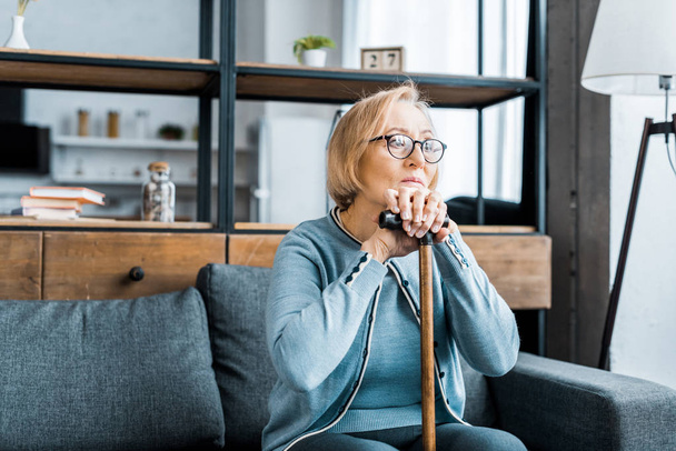 upset senior woman in glasses sitting on couch and leaning on walking stick in living room - Photo, Image