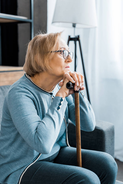 depressed senior woman in glasses sitting with walking stick and looking away at home - Foto, Imagem
