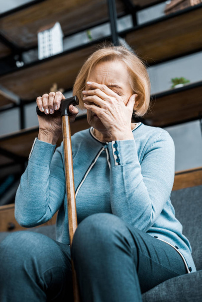 depressed senior woman holding walking stick, covering face with hand and crying at home - Foto, imagen