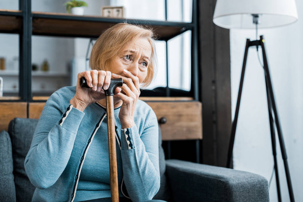 upset senior woman holding walking stick, covering mouth with hand and crying at home with copy space - Fotoğraf, Görsel