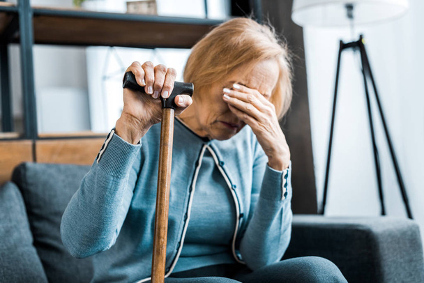 upset senior woman holding walking stick, covering face with hand and crying at home - Photo, Image