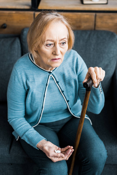 sick senior woman with walking stick sitting on couch and holding medicine in hand  - Fotó, kép