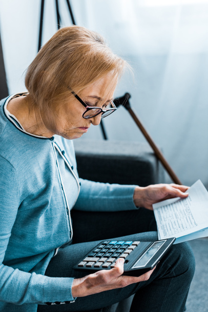 Selektivní fokus starší ženy sedí na gauči s kalkulačkou a počítání bankovek doma - Fotografie, Obrázek