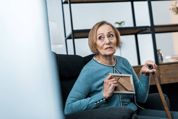zdenerwowany starszy kobieta siedzi z laską i przytrzymanie rama na zdjęcia w domu - Zdjęcie, obraz