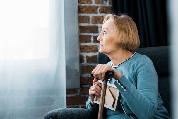 mujer mayor disgustada sentada con bastón y sosteniendo el marco de la foto en casa
 - Foto, Imagen