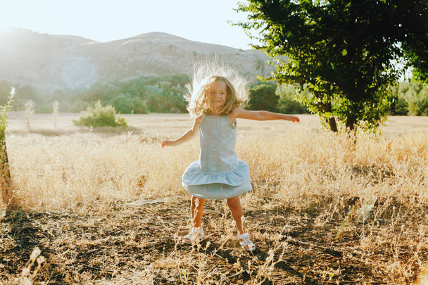μικρό ξανθό κορίτσι με φόρεμα πεδίο με λόφους στο παρασκήνιο - Φωτογραφία, εικόνα