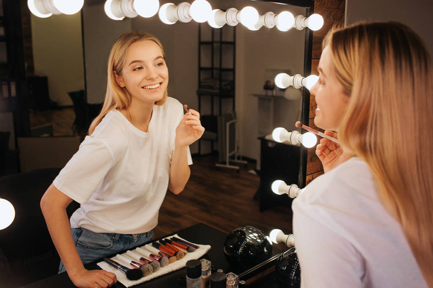 Nice cheerful young woman look in mirror in beauty room and smile. She hold brush for eyeshadows. - Φωτογραφία, εικόνα