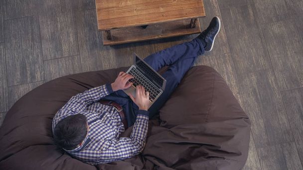 Hombre acostado en un sillón, sosteniendo un portátil y escribiendo
. - Foto, imagen