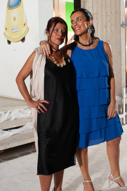 zwei Freundinnen mittleren Alters. eine Frau im Petticoatkleid mit Seitenschlitzen und Spitzeneinsätzen kombiniert mit einer rosa Stola aus Viskose, die andere in einem kleinen blauen Kleid mit Volants. - Foto, Bild