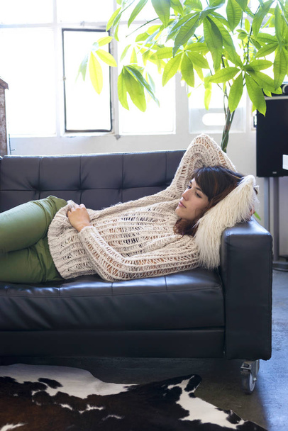 Vista lateral de una hermosa mujer joven tumbada en un sofá de cuero relajándose mientras mira hacia fuera en casa
 - Foto, Imagen
