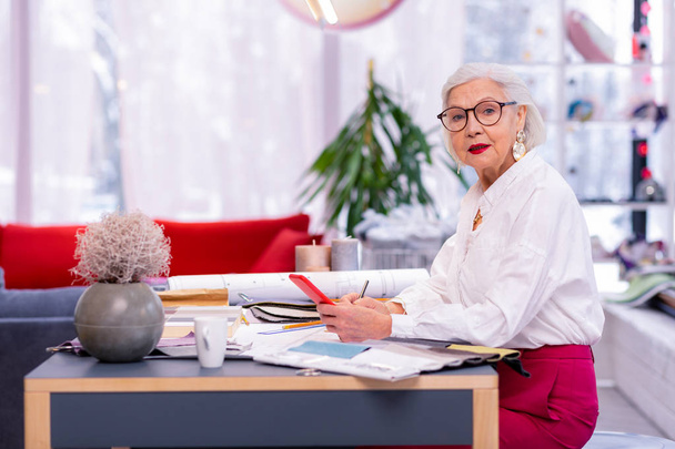 Stylish female executive. Imposing old chief officer in glasses, white shirt and pink trousers holding red phone in hand while sitting at office table - Fotografie, Obrázek