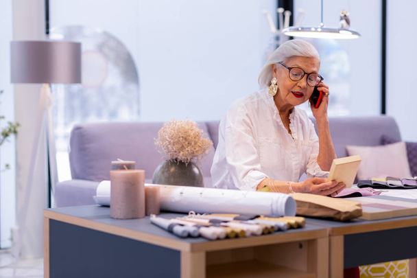 Arranging meeting. Fancy-looking aged female CEO in stylish look and glasses arranging meeting via phone in sophisticatedly furnished office - Foto, Bild