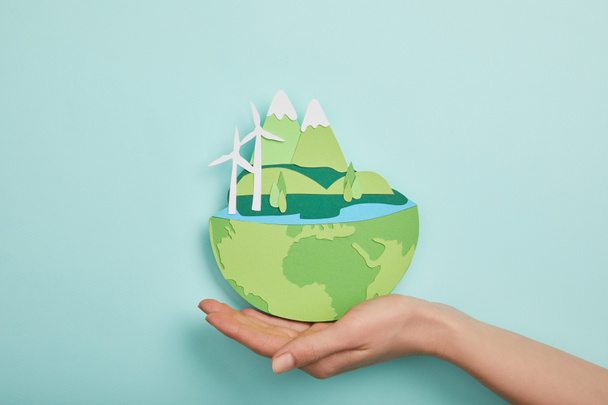 Draufsicht einer Frau, die einen papiergeschnittenen Planeten mit erneuerbaren Energiequellen auf türkisfarbenem Hintergrund hält, Earth Day-Konzept - Foto, Bild
