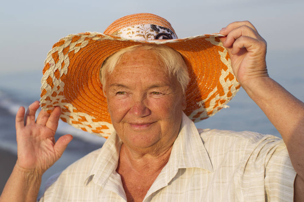 Kaunis iäkäs nainen hatussa meren taustalla. Hymyilevän vanhan naisen kasvot. Mummi lomalla
 - Valokuva, kuva