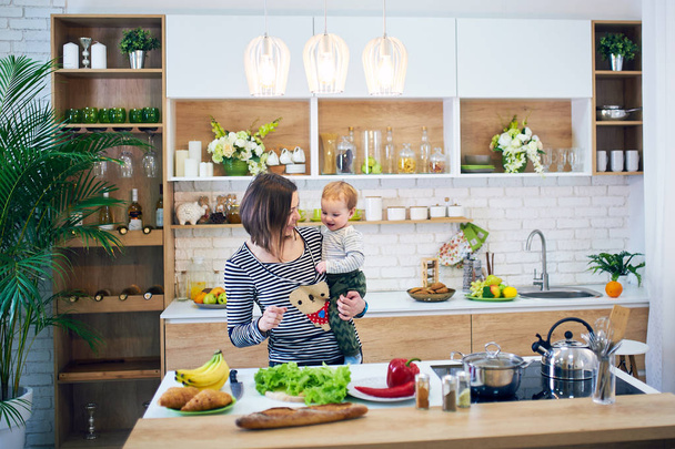 Happy young woman holding a 1 year old child and cooking together in the kitchen - Foto, Bild