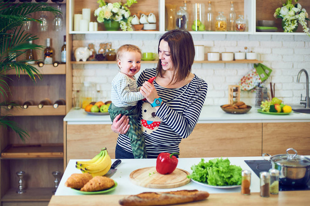 Happy young woman holding a 1 year old child and cooking together in the kitchen - Foto, Bild