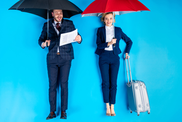 Mulher com valise e homem com jornal de pé sob guarda-chuvas em fundo azul
 - Foto, Imagem