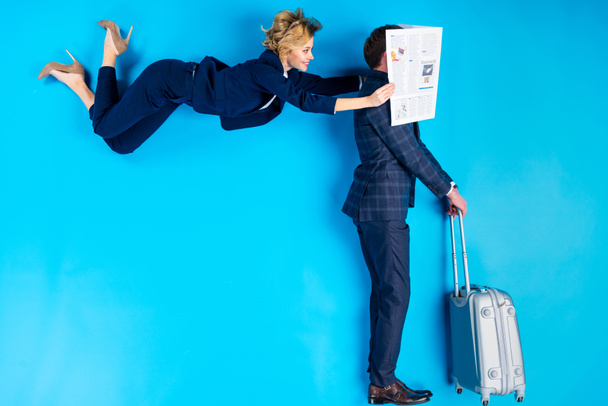Woman posing behind man with suitcase and holding newspaper near his face on blue background - Photo, Image