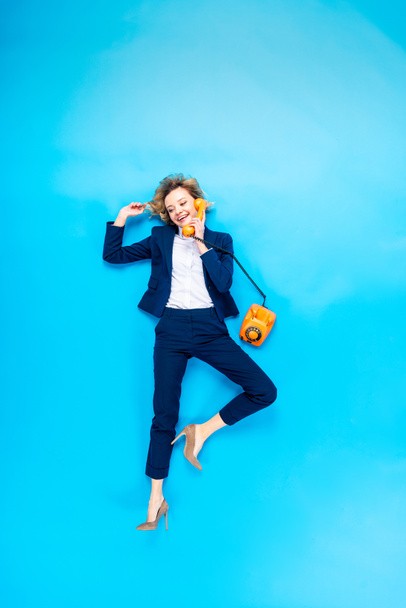 Elegant woman in suit and high-heeled shoes talking on telephone on blue background - 写真・画像