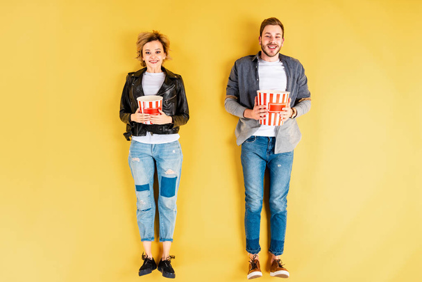 Smiling couple in jeans holding popcorn on yellow background - Foto, Imagen