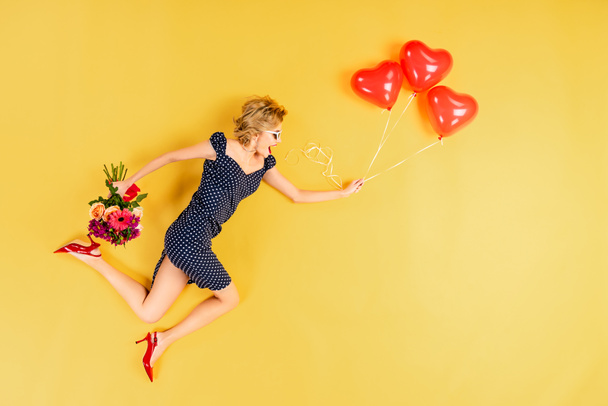 Opgewonden vrouw met lucht Hartvormige ballonnen en bloemen springen op gele achtergrond - Foto, afbeelding