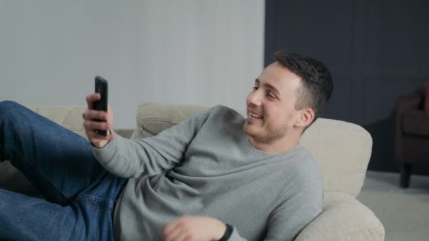 Man at Home Lying on Sofa Does Video Call on Smartphone - Materiał filmowy, wideo