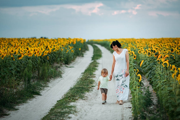 Mutter und Sohn spielen im Sonnenuntergang auf dem Sonnenblumenfeld. sie gehen auf die Straße in der Nähe des Sonnenblumenfeldes - Foto, Bild