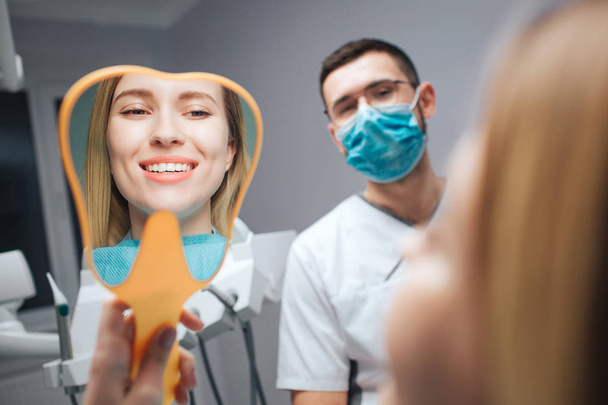 Satisfied female client sit in chair in dentistry room. She look in mirror and smile. Beautiful white teeth. Dentist in mask look at her. - Zdjęcie, obraz