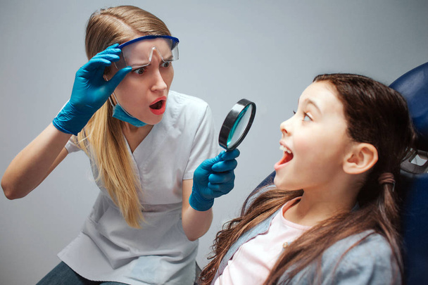 Amazed young female dentist look at girls front teeth through lenz. She is scared. Girl sit in dental chair and keep mouth opened. - Foto, immagini