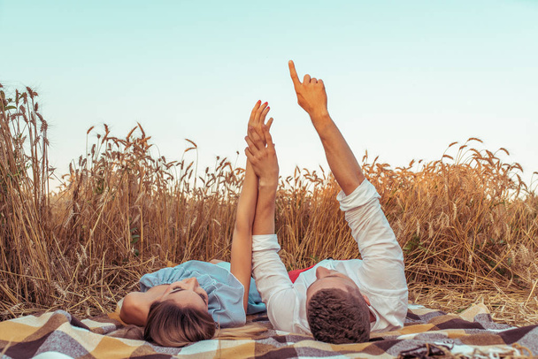 A young couple, man and woman meet sunset and dawn in summer in a wheat field. Lie on bedspread on the ground. Concept of love caring, hugging family happiness and support. Hands show the stars. - Φωτογραφία, εικόνα