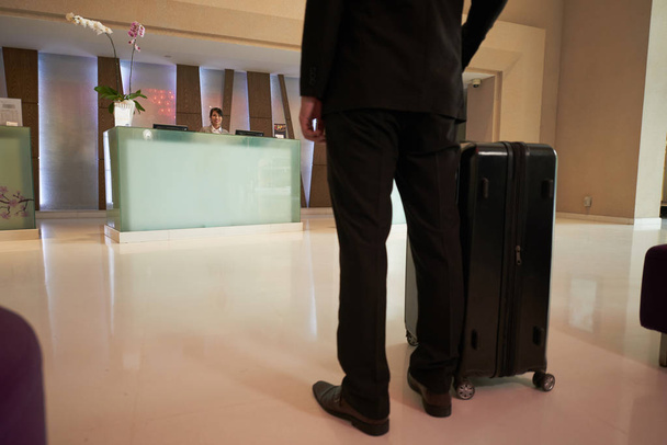 Вид на бизнесмена в черном костюме, стоящего в вестибюле отеля с чемоданом
  - Фото, изображение
