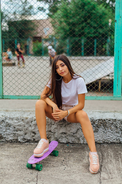 Summer lifestyle image of trendy pretty young girl sitting next to the skateboard coart with her purple plastic skateboard. - Φωτογραφία, εικόνα
