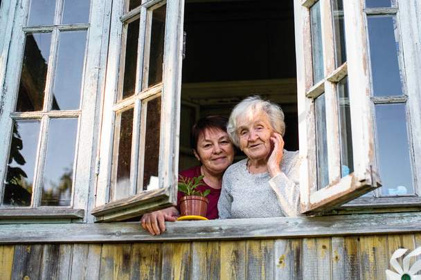Ηλικιωμένη γυναίκα και την κόρη της ενηλίκων από το παράθυρο του σπιτιού του χωριού. - Φωτογραφία, εικόνα