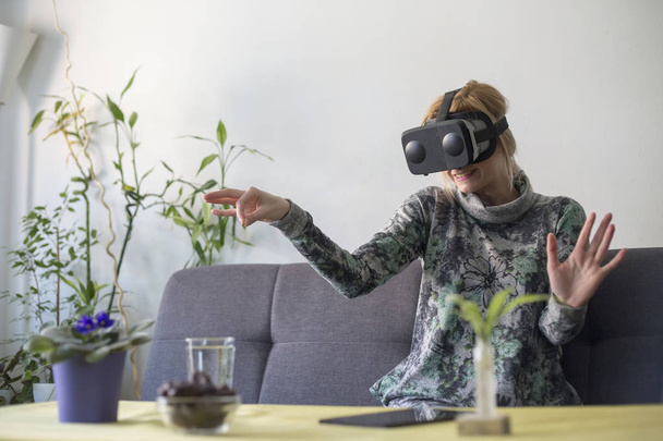 Kobieta w domu przestraszony przy użyciu okulary wirtualnej rzeczywistości na kanapie patrząc wokół i za pomocą gestów z rąk - Zdjęcie, obraz