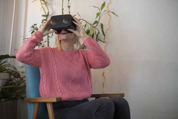 mujer en suéter rosa en casa disfrutando usando gafas de realidad virtual en sillón moderno Mirando a su alrededor y utilizando gestos con las manos
 - Foto, imagen